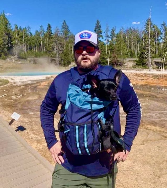 A man wearing a hat and holding his dog in a backpack.